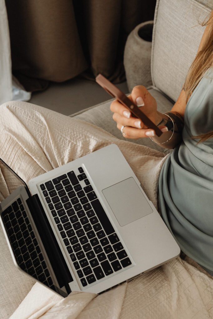 A person is sitting on a beige couch, taking ownership of their life with a smartphone in one hand while a laptop rests on their lap. They wear a light green top and cream-colored pants. The background features a curtain and gray cushion, conveying a relaxed, multitasking environment.