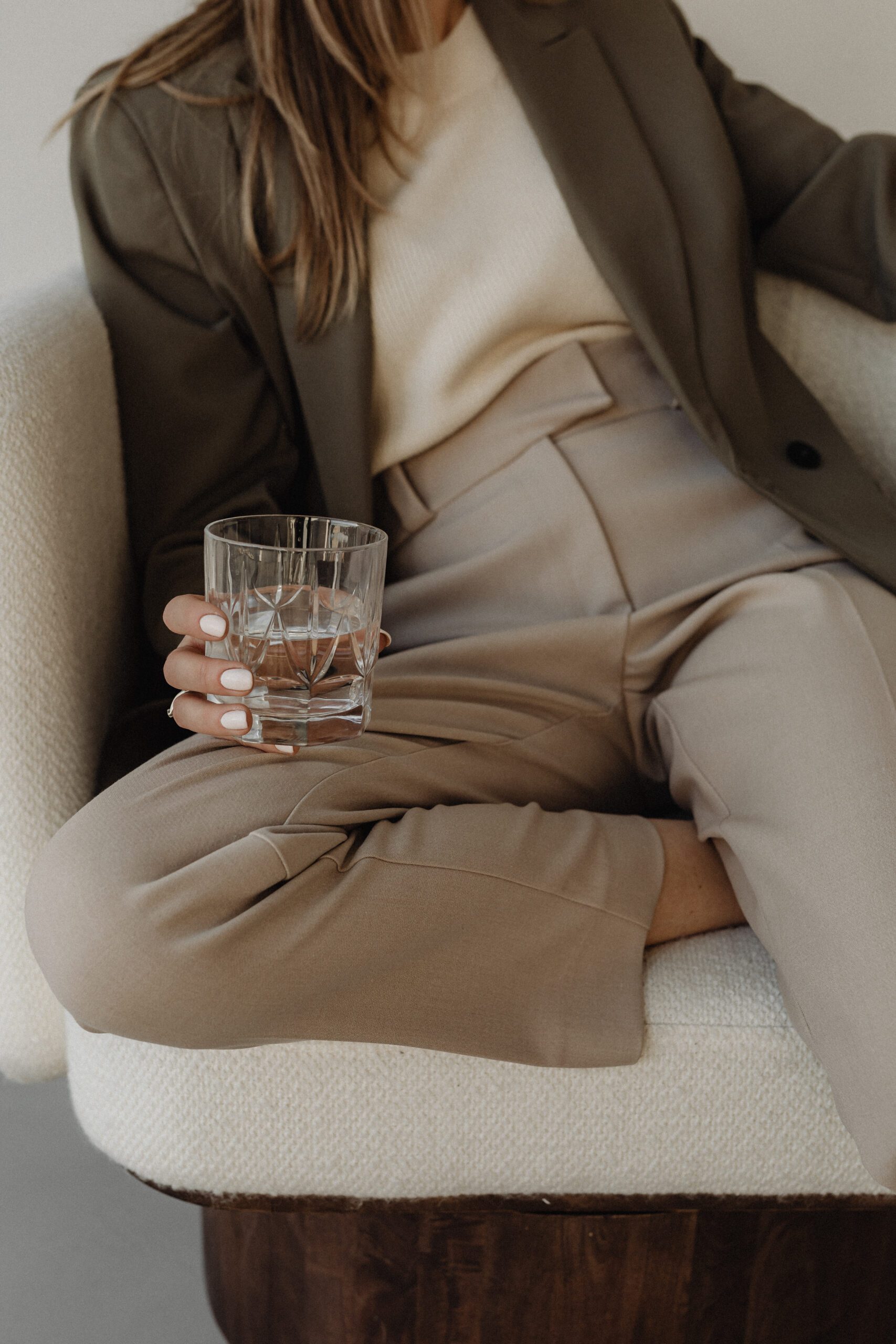 A person elevates their style while sitting on a white chair, holding a glass of water. They wear a beige top, taupe trousers, and an olive-green blazer. Their nails are painted white, and their hair is long and loose. The setting appears calm and minimalistic.