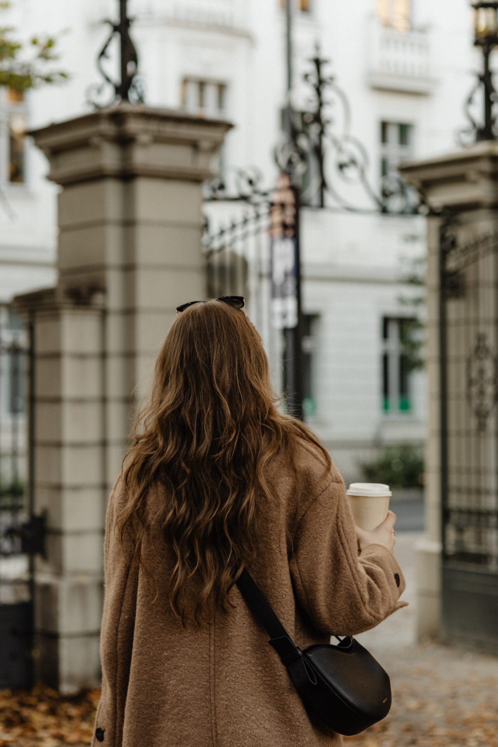 A woman with long, wavy brown hair, wearing a tan coat and carrying a black bag, holds a to-go coffee cup in her left hand. She stands in front of a decorative wrought iron gate and light-colored building, surrounded by fallen leaves. The scene suggests a cool, autumn day.