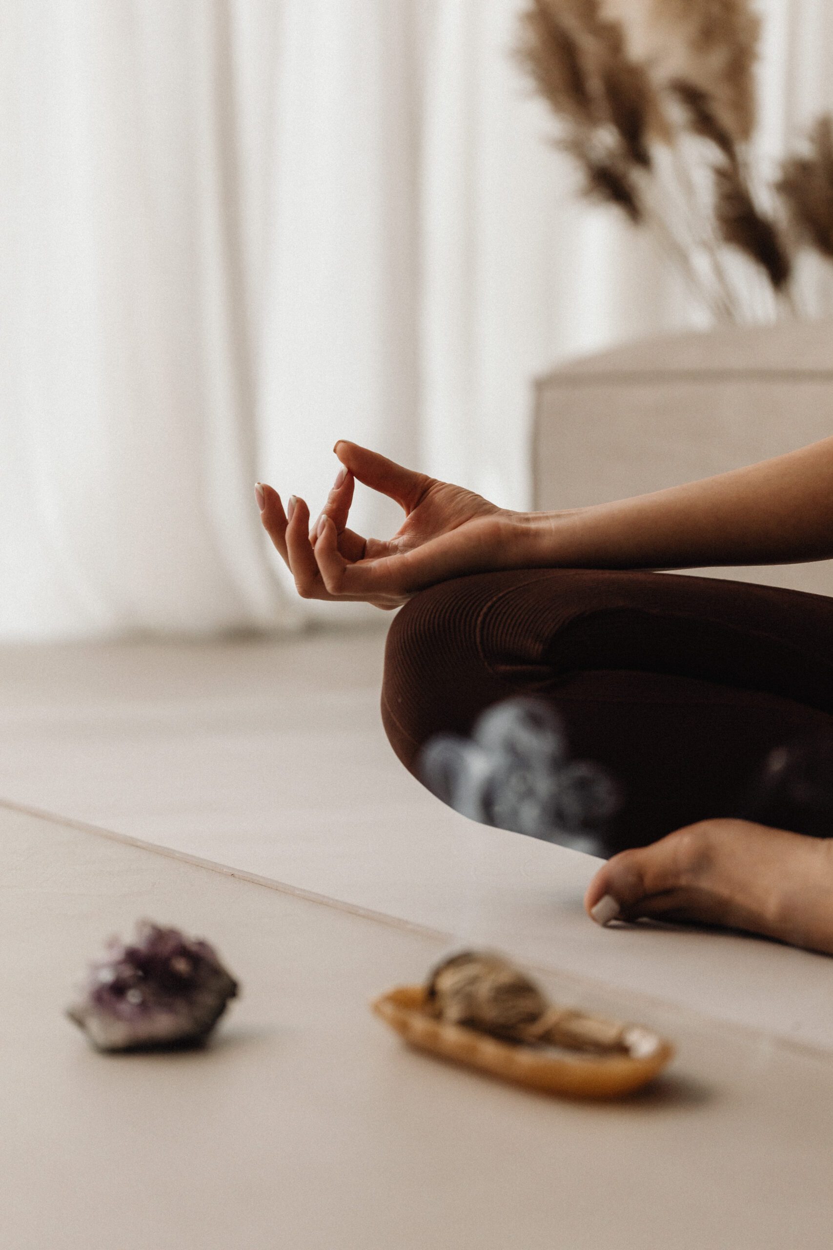 A person is sitting cross-legged on a light-colored floor, focusing on meditation. Their right hand is in a Gyan mudra position, resting on their knee. In the foreground, crystals and a small smudge stick on a tray are visible. Soft, neutral tones and pampas grass in the background create a calming ambiance.