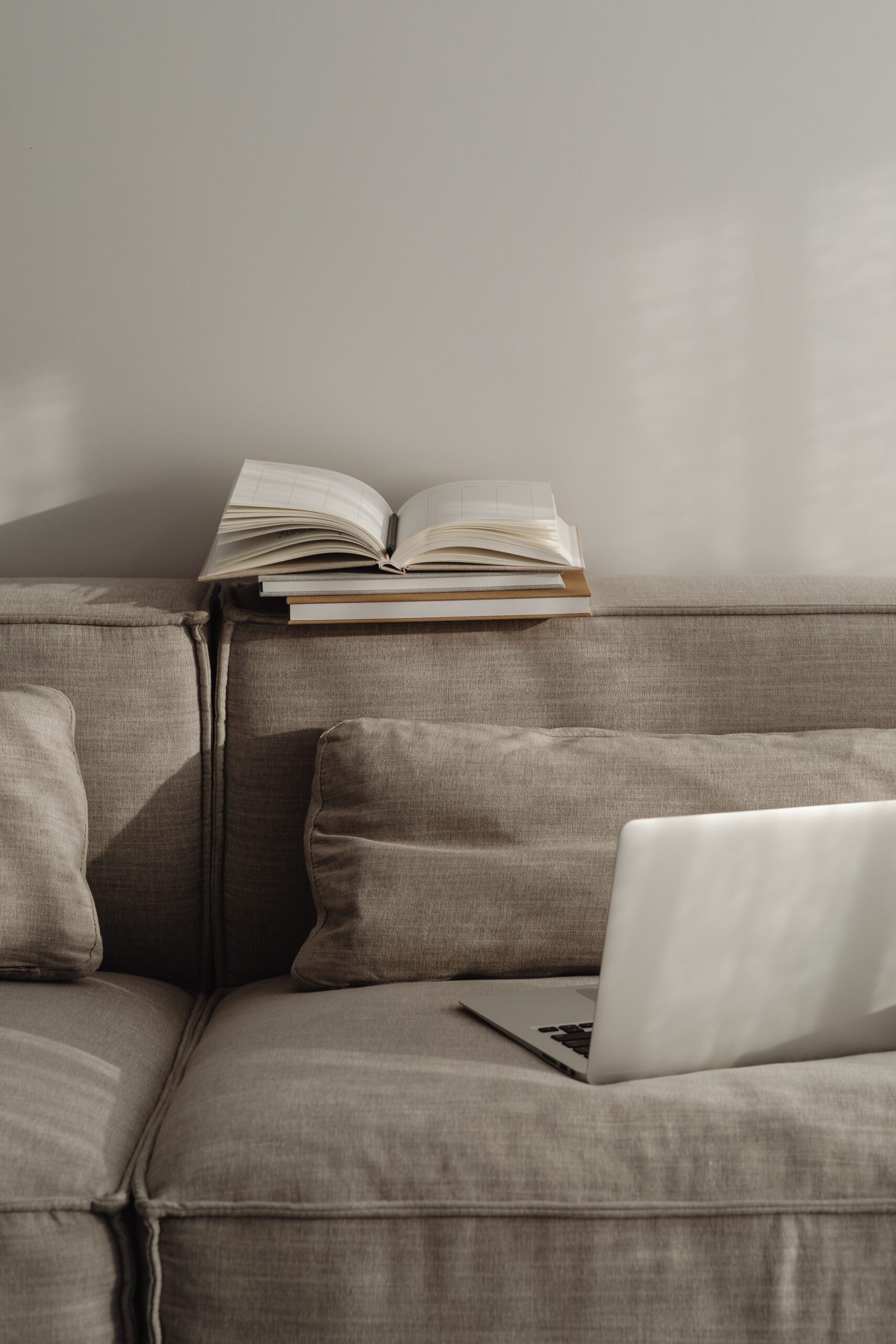 A soft, beige sofa with a couple of plush cushions is in a cozy, sunlit room. An open book rests on the back of the sofa, and a laptop sits on one cushion. Light enters through a window, casting gentle shadows on the wall and furniture, creating a peaceful atmosphere.