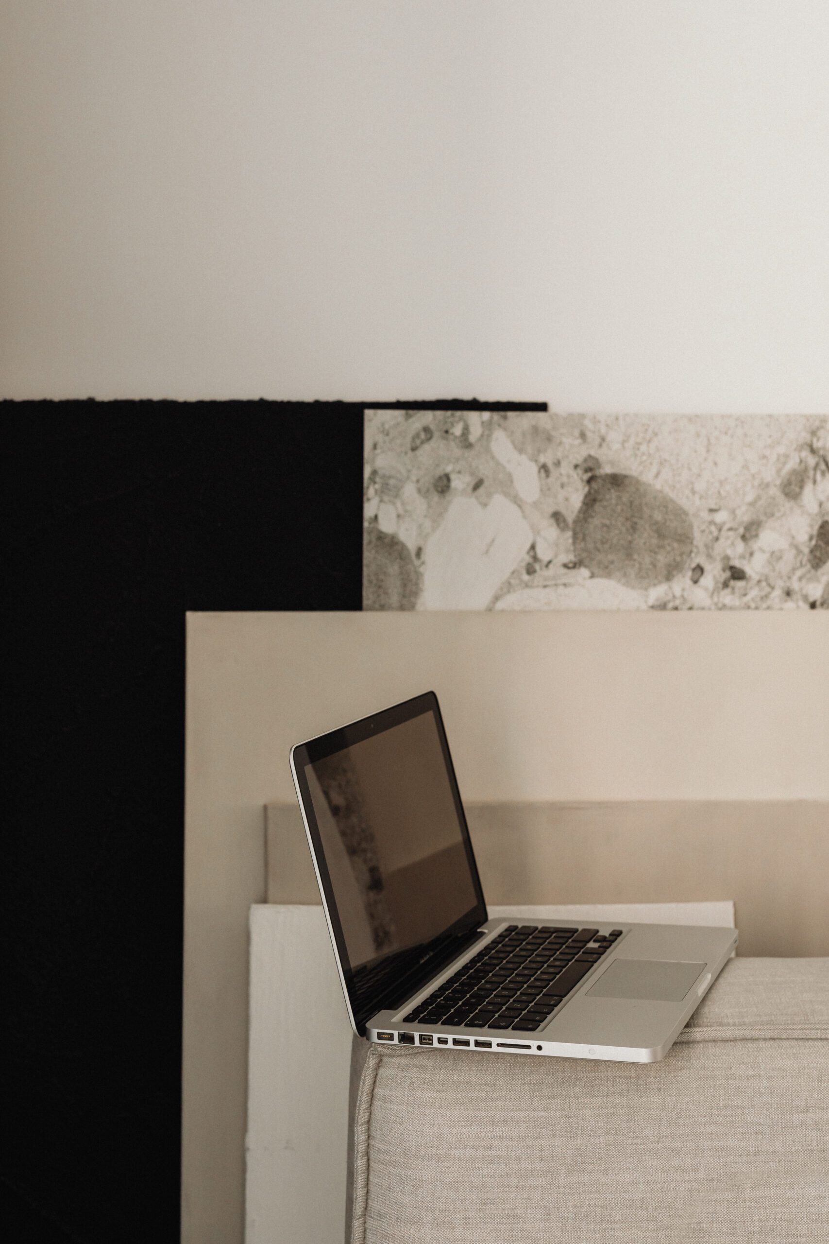 A laptop with a black keyboard and screen is open and resting on a light gray fabric surface. Behind it are textured panels in shades of gray and black, creating a minimalist, modern aesthetic. The background is composed of layered, abstract patterns.