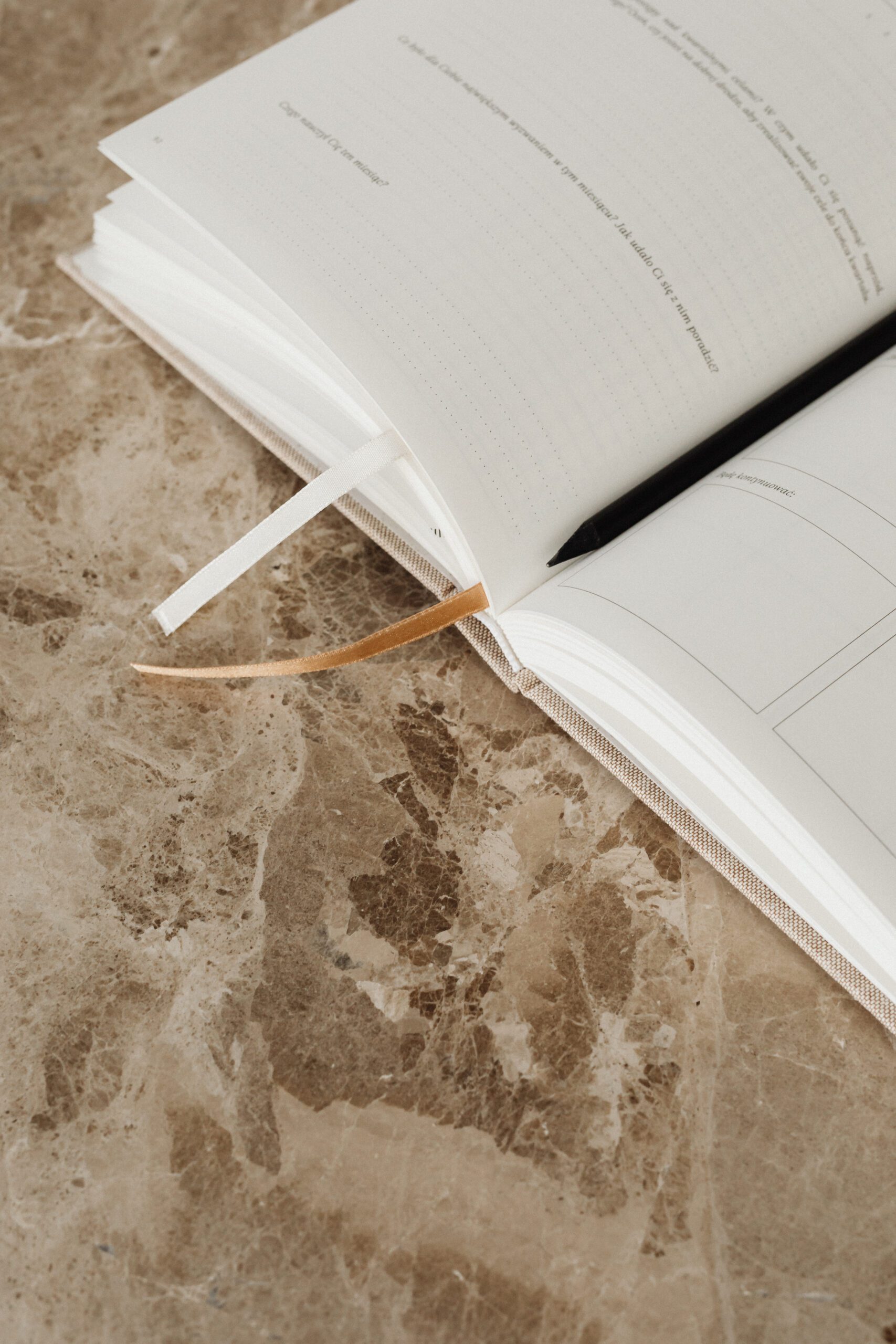 An open notebook with dotted pages and two ribbon bookmarks lies on a textured, brown marble surface. The notebook is positioned towards the upper left corner, with the right side partially visible. The marble surface has a mix of light and dark brown patterns.