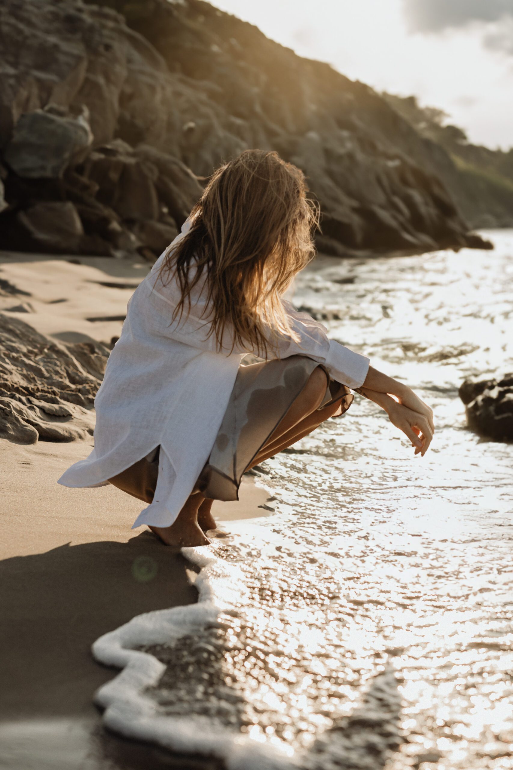 A woman with long hair crouches by the edge of the sea, wearing a white shirt and partially submerged skirt. She touches the water, with rocky cliffs behind her. The sun is low, casting a warm glow over the scene, and gentle waves lap at the shore.