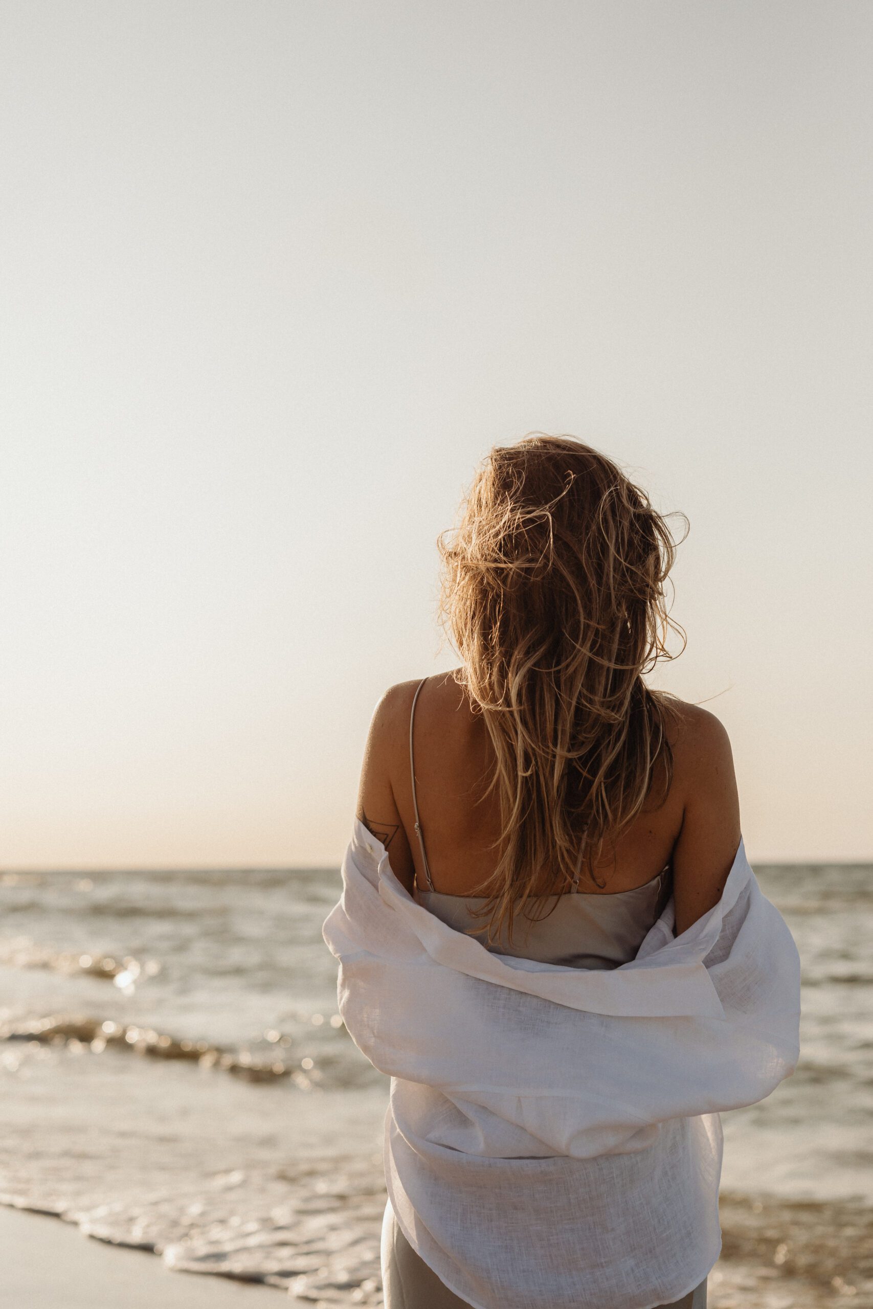 A woman with long hair stands on a beach, facing the ocean. She wears a light dress and a white shirt draped over her shoulders. The sun is setting, casting a warm glow over the scene, and gentle waves ripple in the background. The sky is clear with a soft gradient from blue to orange.