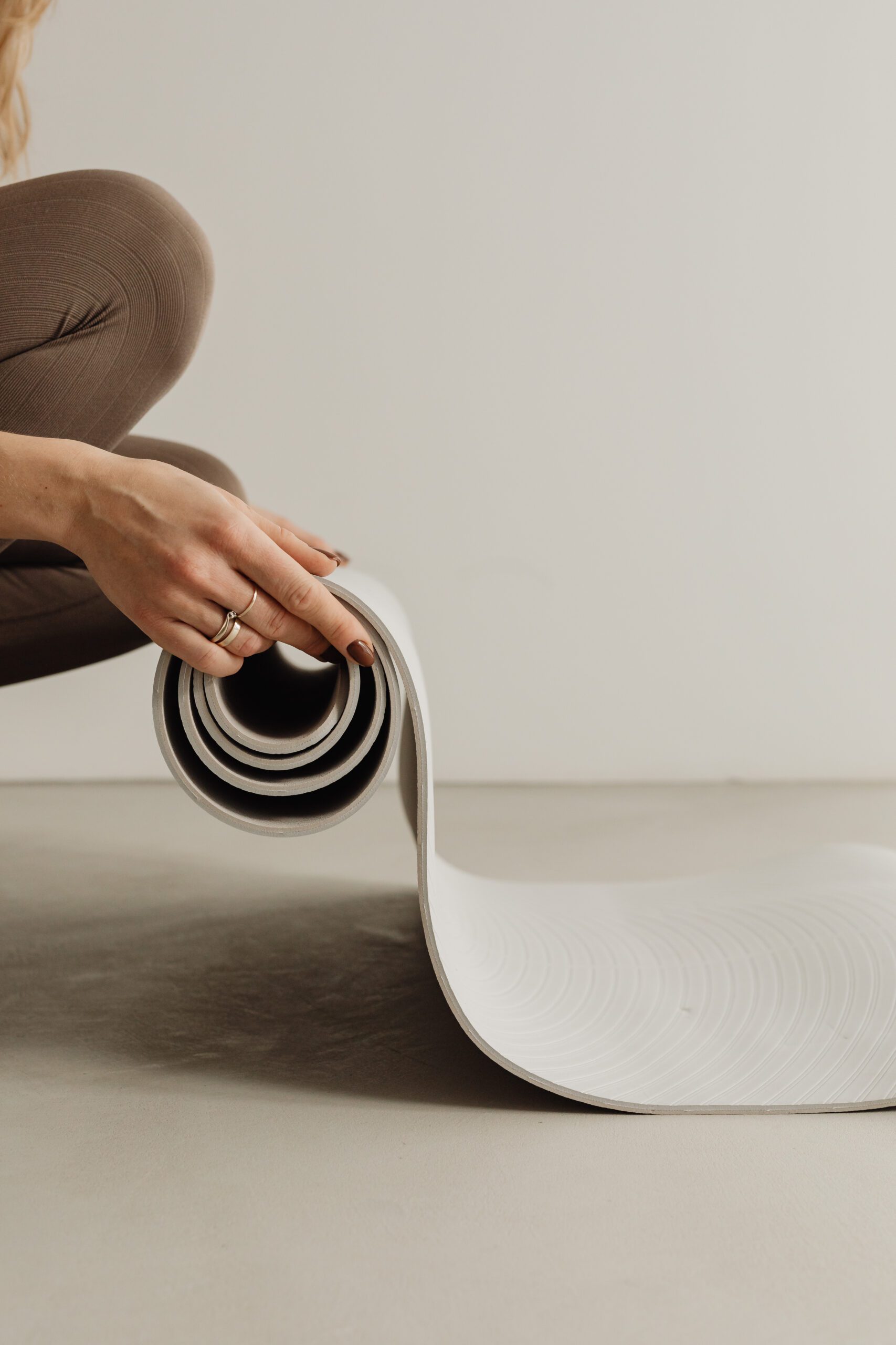 A person in brown pants kneels on the floor, unrolling a white yoga mat. The mat's textured surface is visible as it unfurls. The person's hand, wearing a ring, is holding the mat, against a neutral-toned background. The scene conveys a sense of calm and preparation for exercise or meditation.