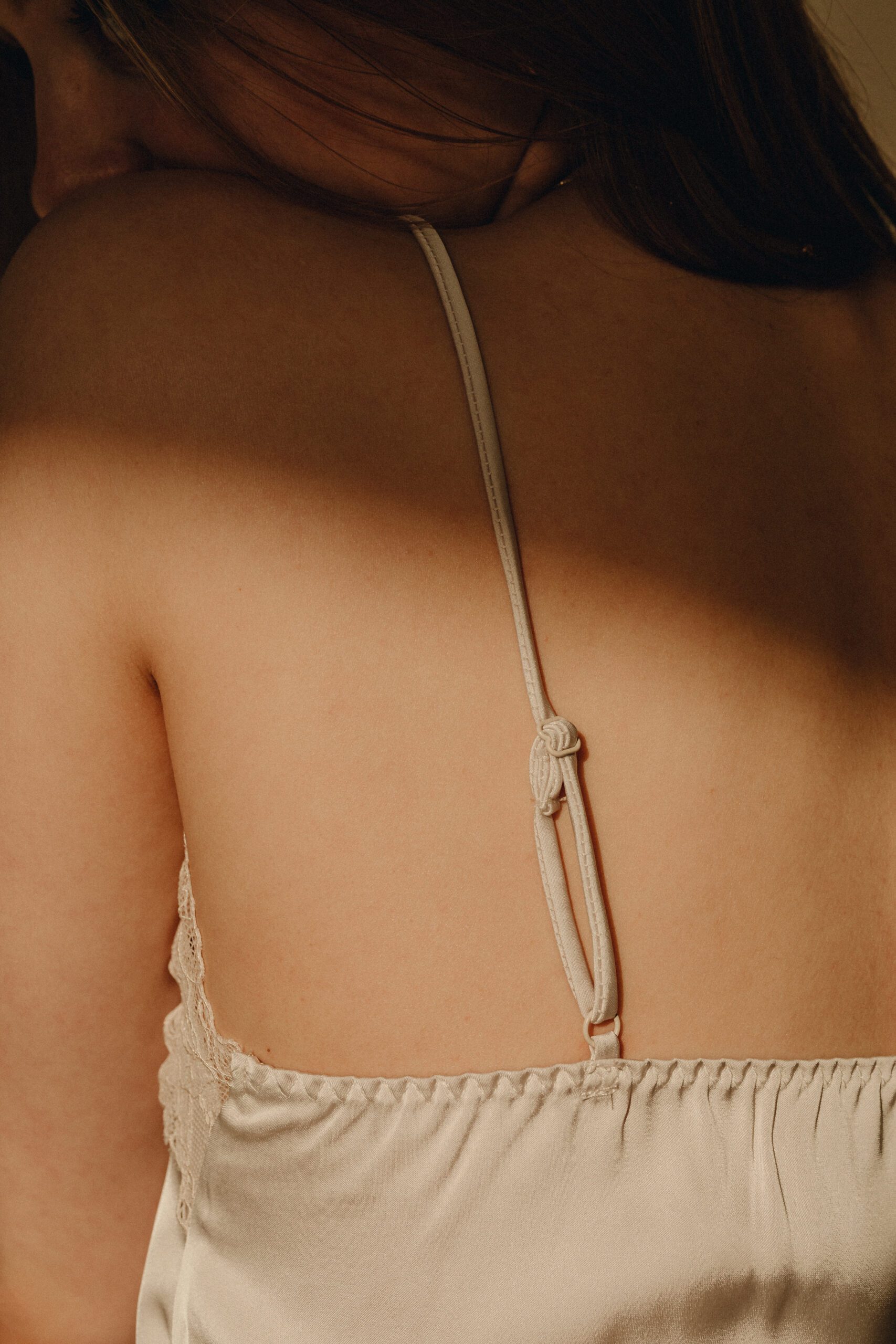 A woman with long hair crouches by the edge of the sea, wearing a white shirt and partially submerged skirt. She touches the water, with rocky cliffs behind her. The sun is low, casting a warm glow over the scene, and gentle waves lap at the shore.