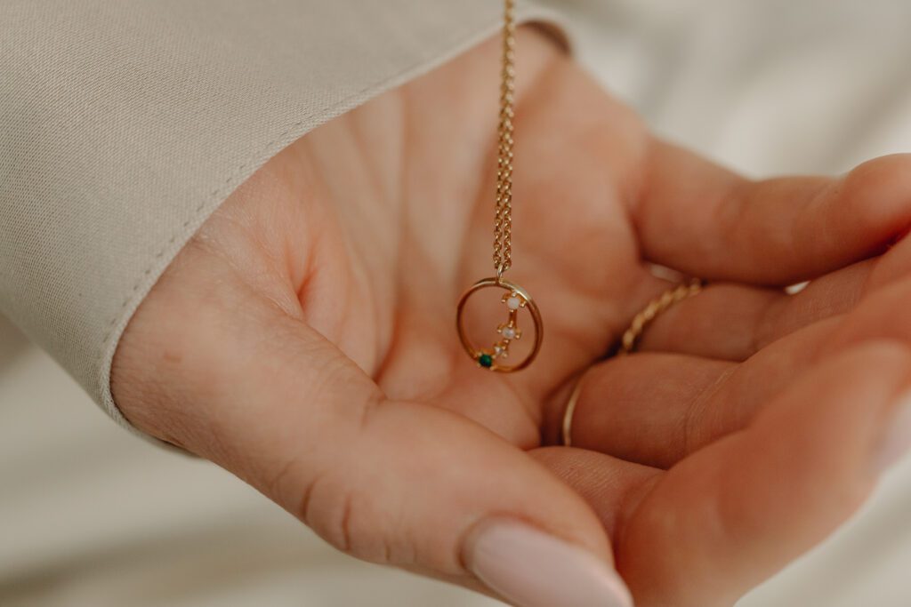 Close-up of a person holding a delicate gold necklace in their palm. The necklace features a circular pendant with small green and clear gemstones, perfect to elevate your style. The person's hand has a pale manicure, and they are wearing a light beige sleeve.
