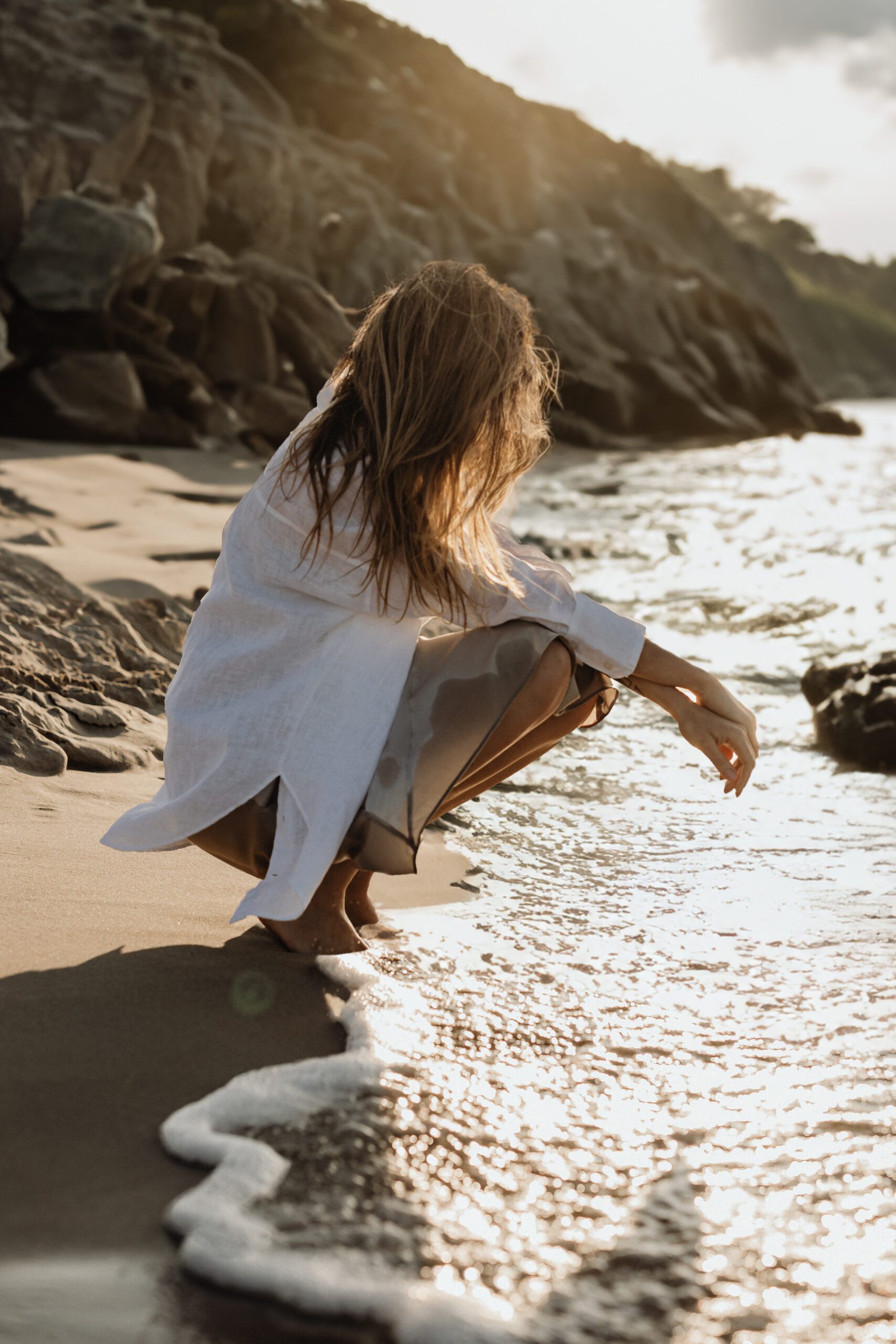 kaboompics_rays-at-the-beach-a-sparkling-moment-32852 A woman with long hair crouches by the edge of the sea, wearing a white shirt and partially submerged skirt. She touches the water, with rocky cliffs behind her. The sun is low, casting a warm glow over the scene, and gentle waves lap at the shore.
