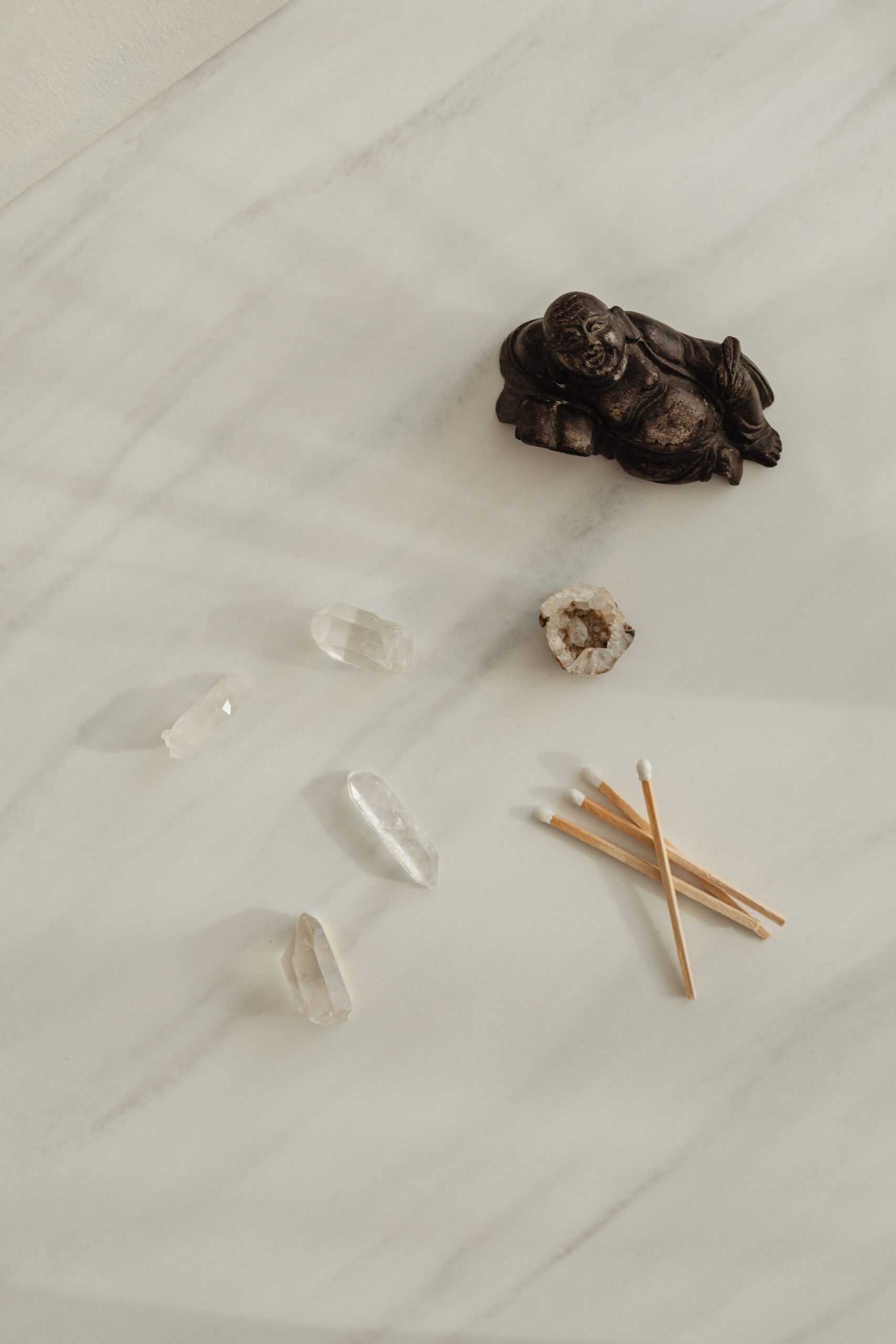 A marble surface displays a small laughing Buddha statue, six clear quartz crystals of varying sizes, an open geode with a crystal center, and four cotton swabs arranged in a neat bundle. The arrangement suggests a calm, meditative atmosphere.