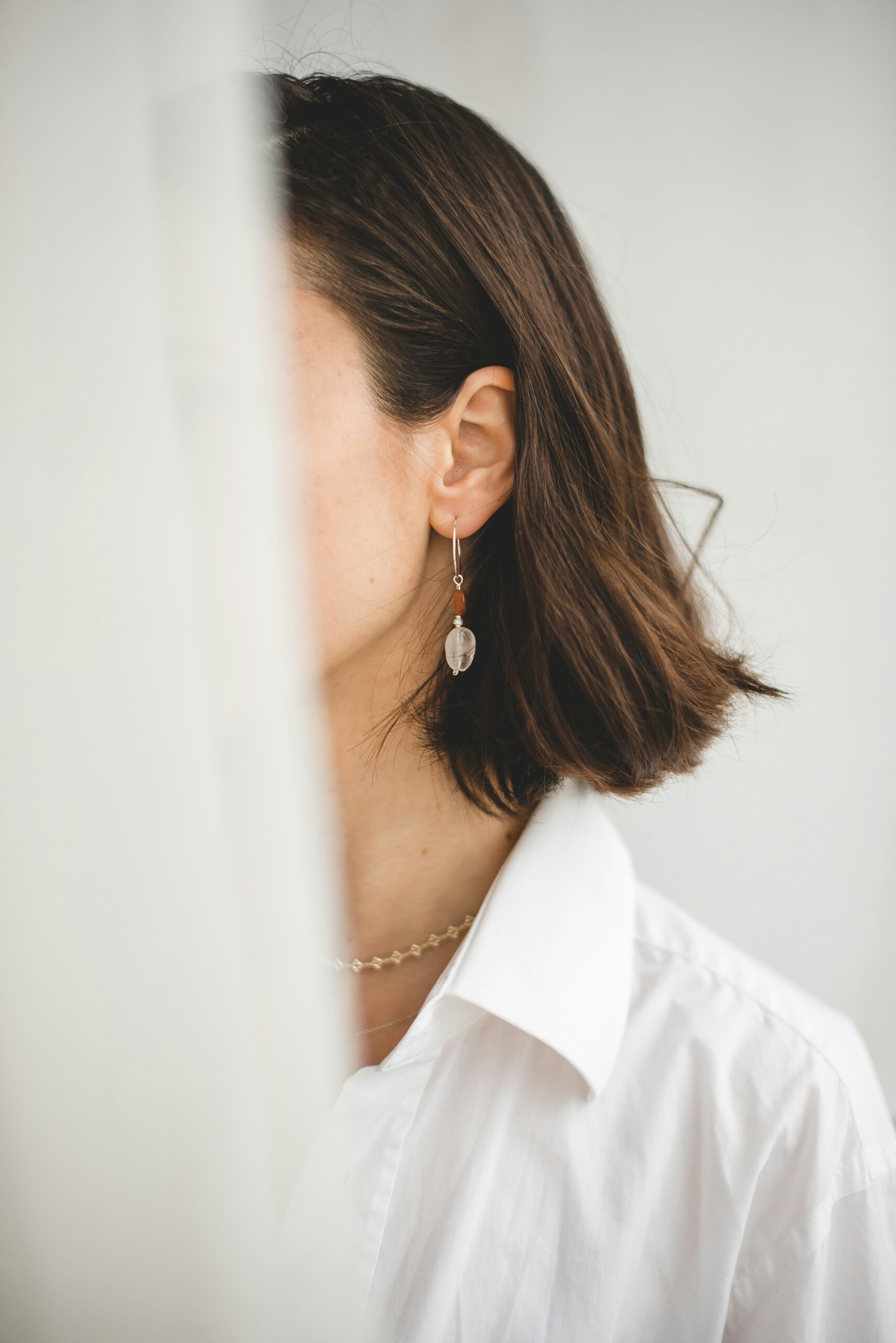 A person with short brown hair is partially obscured by a sheer curtain. They are wearing a white collared shirt and a delicate chain necklace. A dangling earring with a small, translucent pendant is visible. The background is softly blurred, creating a serene atmosphere.