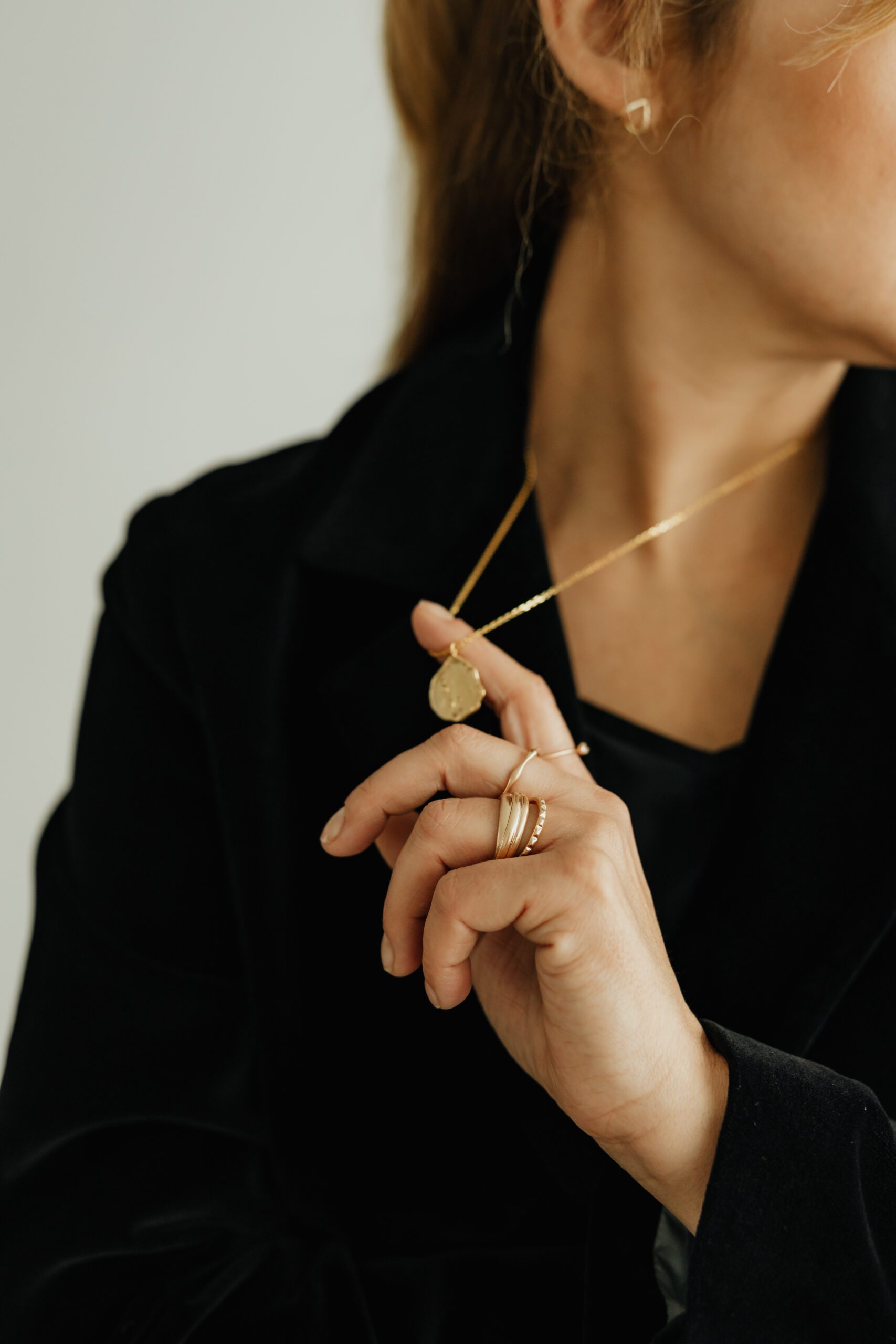kaboompics_jewelry-photo-shoot-woman-wearing-gold-rings-and-necklace-29247 A person with short brown hair is partially obscured by a sheer curtain. They are wearing a white collared shirt and a delicate chain necklace. A dangling earring with a small, translucent pendant is visible. The background is softly blurred, creating a serene atmosphere.
