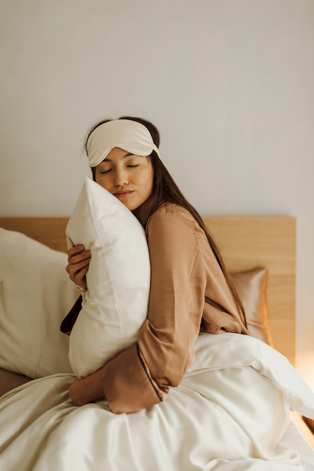 A woman with long hair stands on a beach, facing the ocean. She wears a light dress and a white shirt draped over her shoulders. The sun is setting, casting a warm glow over the scene, and gentle waves ripple in the background. The sky is clear with a soft gradient from blue to orange.